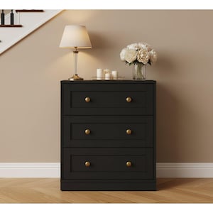 May include: A black three-drawer chest with gold-colored knobs. A lamp with a beige lampshade and a glass vase with white flowers sit on top. The chest is against a beige wall with a staircase in the background.