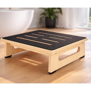 May include: A wooden step stool with a black, textured top and light-colored legs. The top has several parallel cut-out slots. The stool is designed with a rectangular shape and is likely used for reaching higher places.