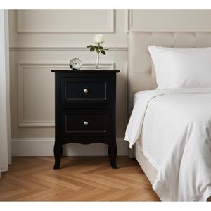 May include: A black two-drawer nightstand with silver knobs, a small clock, and a white rose in a glass vase. The nightstand is next to a bed with white bedding.