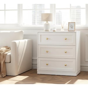 May include: A white three-drawer dresser with gold-colored knobs. A white lamp, a small framed picture, and a diffuser sit on top of the dresser. A white sofa and a tan throw blanket are in the background.