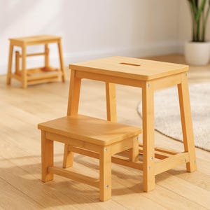 May include: A two-step wooden step stool with a natural finish. The stool has a rectangular top with a handle cutout and a lower step. A smaller, single-step stool is in the background.