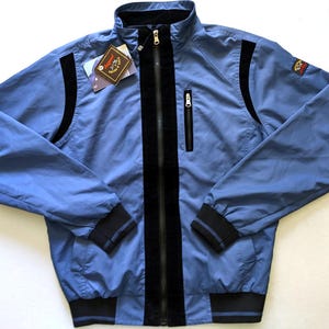 Puede incluir: Chaqueta azul claro con cremallera negra, rayas negras en la parte delantera y detalles negros en las mangas. La chaqueta tiene un pequeño bolsillo en el pecho y una etiqueta adjunta. Cuello alto y puño a rayas negro y azul.