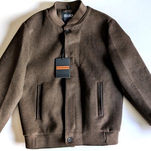 Puede incluir: Una chaqueta bomber marrón con cuello, puños y dobladillo acanalados. La chaqueta tiene cierre frontal de botones, dos bolsillos laterales con ribete oscuro, una etiqueta de la marca Zegna en el cuello y una etiqueta rectangular con rayas naranjas.