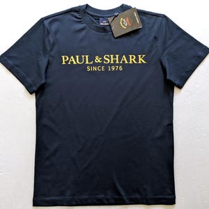 May include: Navy blue t-shirt with the text "PAUL & SHARK SINCE 1976" in gold lettering. The shirt has a crew neck and short sleeves. A tag is attached to the shirt.