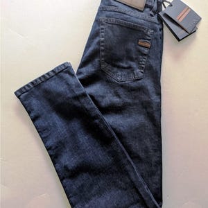 May include: Dark blue denim jeans with a leather patch and a tag. The jeans have a classic five-pocket design and a straight leg fit. The leather patch on the back of the jeans has the brand name "ZEGNA" embossed on it.