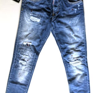 Könnte beinhalten: Blaue Jeans aus Denim mit Used-Look, Rissen, ausgefransten Stellen und verstreuten weißen sowie dunkelroten Farbspritzern. Die Jeans verfügt über einen Knopfverschluss, fünf Taschen und ein schwarzes Markenetikett am Hosenbund.