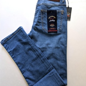 May include: Light blue denim jeans with a classic five-pocket design. The jeans feature a leather patch with a logo and a hang tag. The jeans are folded neatly, showcasing the back pockets and the brand label.