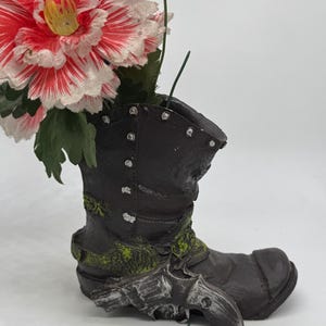 以下が含まれることがあります： 銀色のリボルバーが付いた、装飾用の濃い茶色のブーツ型の花瓶。ブーツには緑色のアクセントがあり、大きな赤と白の花が詰められています。花は黄色の中心と緑の葉を持っています。