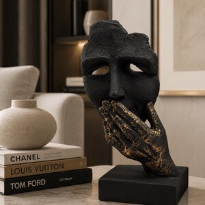 Könnte beinhalten: Eine schwarze Skulptur eines Gesichts mit einer Hand, die den Mund bedeckt. Die Hand hat eine goldfarbene Oberfläche. Die Skulptur steht auf einem schwarzen Sockel. Dekorative Bücher und eine Vase befinden sich im Hintergrund.