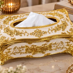 May include: A rectangular tissue box holder featuring a white base with ornate gold-colored floral relief patterns and decorative accents. The top surface includes a central opening with a white tissue protruding, and the base rests on four small feet.