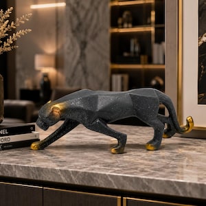Puede incluir: Una figura de pantera negra con detalles dorados en las patas, orejas y cola. El diseño geométrico está colocado sobre una superficie con efecto mármol. Un jarrón con plantas secas y libros se encuentran en el fondo.