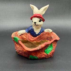 Könnte beinhalten: Ein skurriler Keramik-Blumentopf mit einem weißen Hasen, der einen roten Hut und ein blaues Hemd trägt. Der Hase befindet sich in einem strukturierten, rötlich-orangen Topf mit grünen Akzenten, ideal für kleine Pflanzen oder Dekorationen.