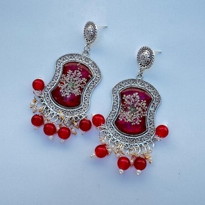 Könnte beinhalten: Ein Paar silberfarbene Hängeohrringe. Jeder Ohrring hat einen rechteckigen, verzierten Rahmen mit einem tiefroten Mittelteil, der mit einem floralen Design verziert ist. Rote Perlen und kleine weiße Perlen hängen von unten.