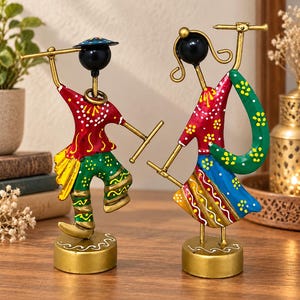 Peut inclure: Deux figurines en métal représentant des danseurs sur des socles dorés circulaires. La figurine de gauche porte un haut rouge et un pantalon vert à motifs jaunes. Celle de droite porte un haut rouge et une jupe bleue avec une écharpe verte.