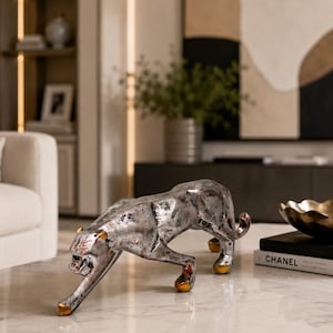Könnte beinhalten: Eine silberfarbene Pantherfigur in schleichender Haltung mit einer strukturierten, abgenutzten Oberfläche. Die Skulptur weist goldfarbene Akzente an den Ohren und Pfoten auf, wobei das Tier in einer tiefen, nach vorne geneigten Position dargestellt ist.