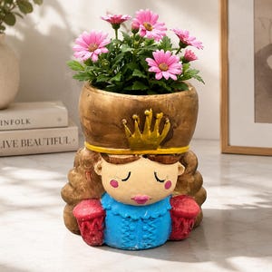 Könnte beinhalten: Ein dekorativer Übertopf in Form einer Figur mit goldener Krone, blauem Oberteil und roten Ärmeln sowie braunem Haar. Die Figur hat geschlossene Augen und rosa Wangen. Aus der Krone wächst ein Büschel leuchtend rosa, gänseblümchenartiger Blumen.