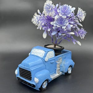 Könnte beinhalten: Ein blauer Vintage-Truck-Pflanztopf mit schwarzem Kühlergrill und silbernen Akzenten. Die Ladefläche des Trucks enthält einen kleinen schwarzen Topf mit lila und weißen Kunstblumen. Der Pflanztopf steht vor einem dunkelgrauen Hintergrund.