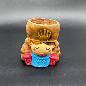 Könnte beinhalten: Ein skurriler Keramik-Blumentopf in Form einer Cartoon-Prinzessin. Der Pflanztopf hat einen holzfarbenen Fasskopf, eine goldene Krone und ein blaues Kleid. Die Prinzessin hat geschlossene Augen, rosige Wangen und rote Akzente. Der Pflanztopf steht auf einer schwarzen Oberfläche.