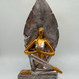 Könnte beinhalten: Yoga-Figur mit einer goldfarbenen Figur in sitzender Pose, Hände zum Gebet gefaltet. Die Figur ist vor einem großen, silberfarbenen Blatt mit roten Akzenten platziert. Der Sockel ist ein strukturierter, rechteckiger Block.