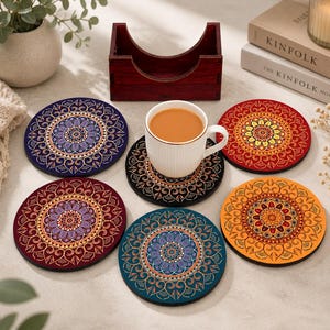 Könnte beinhalten: Ein Set aus sechs runden Untersetzern mit komplexen Mandala-Mustern in verschiedenen Farben wie Blau, Rot, Orange und Violett, ergänzt durch einen dunklen Holzhalter und eine weiße Keramiktasse mit einem hellbraunen Getränk.
