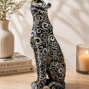 May include: A seated leopard figurine featuring a dark charcoal base color with intricate, raised silver and gold scrollwork patterns covering its entire body. The sculpture is depicted in a poised, upright position with its head tilted slightly upward.