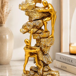 Puede incluir: Una escultura decorativa que presenta dos figuras humanas de color dorado escalando una formación rocosa texturizada en plata y oro. Las figuras están representadas en movimiento, una alcanzando hacia arriba y la otra posicionada más abajo, ambas unidas a la estructura de piedra.