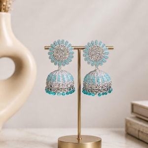 Könnte beinhalten: Ein Paar silberfarbene Jhumka-Ohrringe mit einem runden Stecker und einem glockenförmigen Anhänger, besetzt mit hellblauen Steinen und klaren Kristallen. Der untere Rand jeder Glocke ist mit einer Reihe kleiner, hängender hellblauer Perlen verziert.