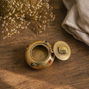 Peut inclure: Un ancien coffret à bijoux en laiton avec un couvercle à charnière, orné de pierres précieuses colorées. La boîte ronde est ouverte, révélant l'intérieur. Le couvercle est rectangulaire avec un petit motif en relief. La boîte est sur une surface en bois.