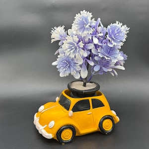 Może przedstawiać: Żółta doniczka w kształcie samochodu z czarnym wnętrzem, w której znajduje się mała roślina z fioletowo-białymi kwiatami. Samochód ma czarne koła i białe akcenty.
