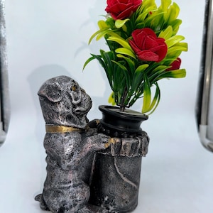 Könnte beinhalten: Dekorative Hundefigur, die einen Blumentopf hält. Der Hund ist silber-grau mit goldenen Akzenten. Der schwarze Blumentopf enthält rote Rosen und grüne und gelbe Blätter.
