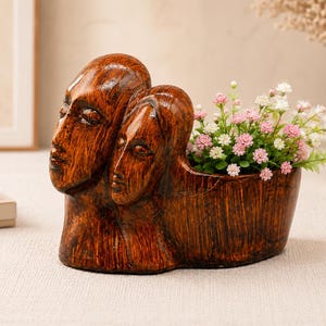 Könnte beinhalten: Ein brauner Pflanztopf mit Holzstruktur, der an der Seite zwei skulptierte menschliche Gesichter zeigt. Der Topf ist mit kleinen künstlichen rosa und weißen Blumen gefüllt. Die Oberfläche hat ein dunkles, poliertes Aussehen mit Streifen.