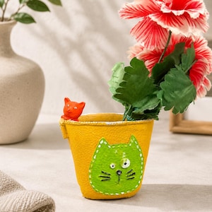 Könnte beinhalten: Ein gelber, eimerförmiger Übertopf mit einer grünen Katzenkopf-Applikation mit gestickten Details und einer kleinen orangefarbenen Katzenfigur am Rand. Der Übertopf enthält rote und weiße Blumen mit grünen Blättern.