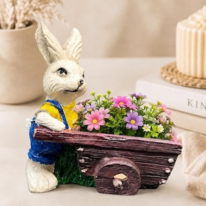 Könnte beinhalten: Eine weiße Hasenfigur mit gelbem Hemd und blauer Latzhose, die hinter einem dunkelbraunen Pflanzgefäß in Form einer Schubkarre steht, gefüllt mit kleinen künstlichen rosa, violetten und weißen Blumen sowie grünem Blattwerk.