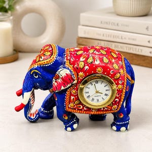 Può includere: Una statuetta di elefante blu con una coperta rossa decorata da motivi floreali dorati e bianchi. Un orologio analogico rotondo con cornice dorata e quadrante bianco con numeri romani neri è incassato sul fianco dell'elefante dalle zanne bianche.