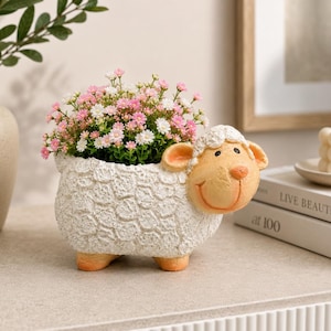 Könnte beinhalten: Ein weißer, schafförmiger Übertopf mit strukturierter, wolleartiger Oberfläche, orangefarbenen Füßen und einem lächelnden Gesicht. Der Übertopf ist mit einem Büschel kleiner rosa und weißer Blumen gefüllt und dient als Dekoration.