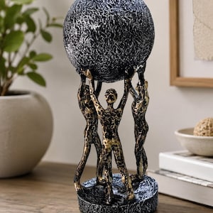 May include: A decorative sculpture featuring three figures standing on a circular base, collectively holding a large, textured sphere above their heads. The piece is finished in a distressed black and gold metallic paint with a rough, uneven surface.
