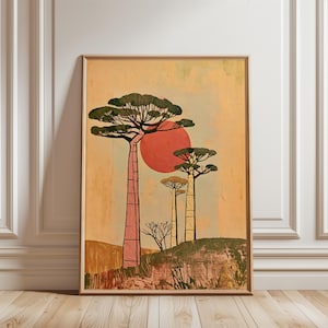 Op de afbeelding: Een ingelijste muurkunstprint met een gestileerd landschap met drie hoge baobabbomen in roze en crèmetinten tegen een gedempte oranje achtergrond. Een grote, effen rode cirkel die de zon voorstelt, bevindt zich achter de bomen boven een getextureerde grond.