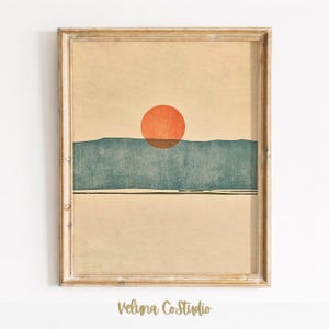 May include: A framed wall art print featuring a textured beige background with a large, solid orange circle positioned above a horizontal, dark teal landscape shape. The artwork is enclosed in a light brown wooden frame with a visible grain.