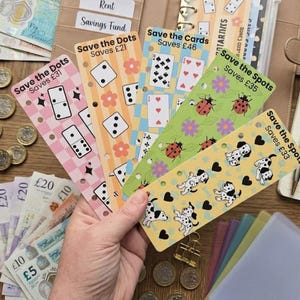 May include: Five rectangular paper savings trackers with punched holes for a binder. The trackers feature colourful patterns including dominoes, dice, playing cards, ladybugs, and dalmatian puppies, with text indicating savings goals ranging from £21 to £46.
