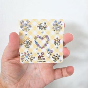 May include: A square savings tracker card with a yellow and white gingham pattern featuring various floral illustrations, a heart-shaped flower wreath, a small mushroom, and a snail. The card includes small circles labeled £1 and the text Saves £10.
