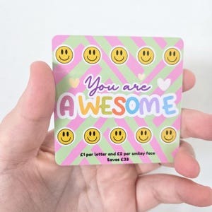 May include: A square card with a light green and pink diagonal striped background featuring yellow smiley faces, small hearts, and the text "You are AWESOME" in colourful letters. The bottom text reads "£1 per letter and £2 per smiley face Saves £33".