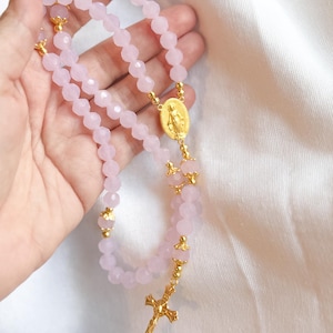 Puede incluir: Un rosario rosa claro con cuentas facetadas y detalles metálicos dorados. El rosario presenta una cruz dorada, un medallón con una figura religiosa y adornos dorados.