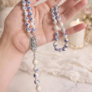 Op de afbeelding: Een rozenkrans met witte en blauwe bloempatroon kralen. De rozenkrans heeft een zilverkleurig middelpunt met een religieuze figuur en een kruisvormige hanger. De kralen zijn geregen met zilverkleurig metaal. Een religieus item.