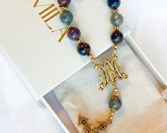 May include: A rosary with multi-colored gemstone beads and gold-colored metal accents. The rosary features a crucifix and a centerpiece with an image. The rosary is presented in a white box with the word "MILA" in gold lettering.