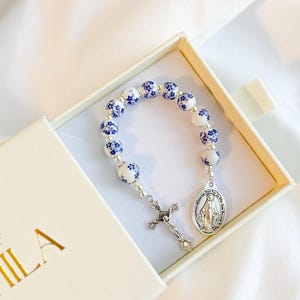 Puede incluir: Un rosario de pulsera con cuentas florales blancas y azules, una cruz y un colgante ovalado plateado. La pulsera se presenta en una caja abierta de color blanco roto con letras doradas. Las cuentas tienen un estampado floral azul.