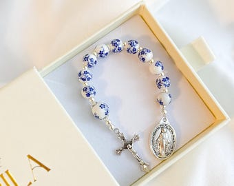 May include: A white and blue floral bead rosary bracelet with a silver crucifix and a silver oval pendant. The bracelet is presented in an open, off-white box with gold lettering. The beads have a blue floral pattern.