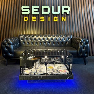 Könnte beinhalten: Ein schwarzes Chesterfield-Ledersofa und ein moderner Glastisch mit schwarzem Marmorsockel. Der Couchtisch ist mit blauen Lichtern beleuchtet und enthält verschiedene Gegenstände. An der Wand hinter dem Sofa steht der Text "SEDUR DESIGN".
