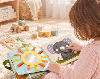 May include: A soft fabric activity book featuring a sun design with a central circular mirror and yellow and orange felt rays on one page, and a grey felt elephant with polka dot ears on the opposite page, designed for tactile sensory play.