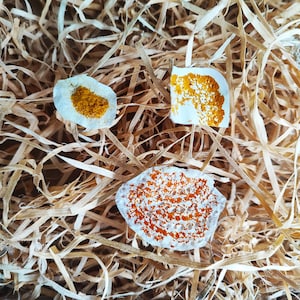 Peut inclure: Trois objets blancs de forme irrégulière avec des surfaces texturées orange et jaune reposent sur un lit de paille brune. Les objets ont une apparence rugueuse et organique, avec les couleurs orange et jaune contrastant avec le blanc.