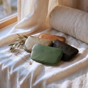 May include: A collection of four small, rectangular leather pouches in shades of olive green, sage green, dark charcoal, and tan, accompanied by a delicate gold chain necklace and a small sprig of green leaves with slender, pointed foliage.